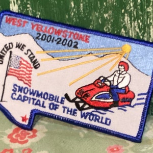 May include: A blue and white embroidered patch with a snowmobile and a sun rising over a mountain. The text on the patch reads "West Yellowstone 2001-2002", "United We Stand", and "Snowmobile Capital of the World".