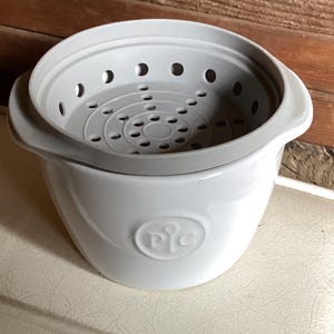 May include: A white ceramic steamer pot with a gray, perforated insert. The pot has two handles and a rounded shape. The embossed logo "PIC" is visible on the front. The steamer is designed for cooking food.