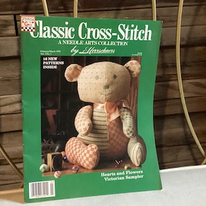 May include: A green magazine cover with the title "Classic Cross-Stitch" and a picture of a cross-stitch pattern of a teddy bear. The magazine is from the Needle Arts Collection by Herrschners. The text "Hearts and Flowers Victorian Sampler" is at the bottom of the cover.
