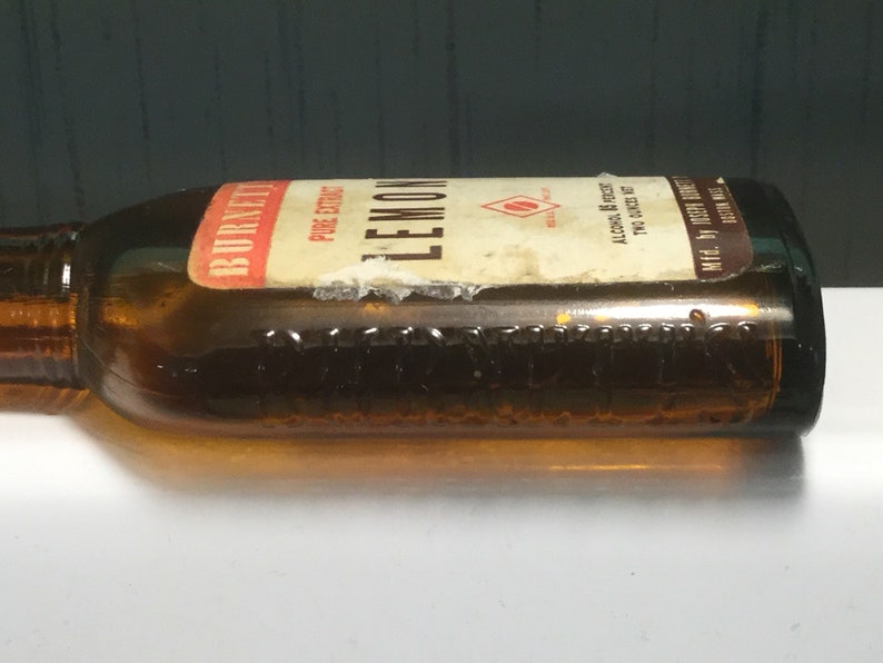 May include: A brown glass bottle with a label that reads "Burnett's Lemon Pure Extract" and "Alcohol 6% by Volume 70% Alcohol by Weight".