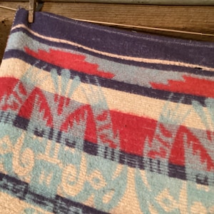 May include: A woven blanket with a geometric pattern in red, blue, and light blue. The blanket is made of a soft material and has a dark blue border. The design features repeating shapes and lines.