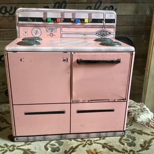 May include: A vintage pink toy stove with a black handle and a WOLVERINE logo. The stove has four burners, an oven, and two drawers.