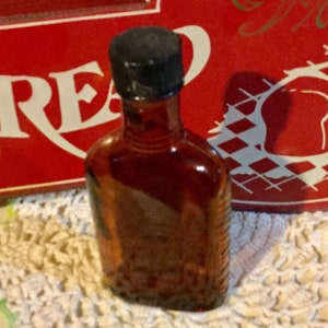 May include: A small, amber-colored glass bottle with a black cap. The bottle is sitting on a white crocheted doily.