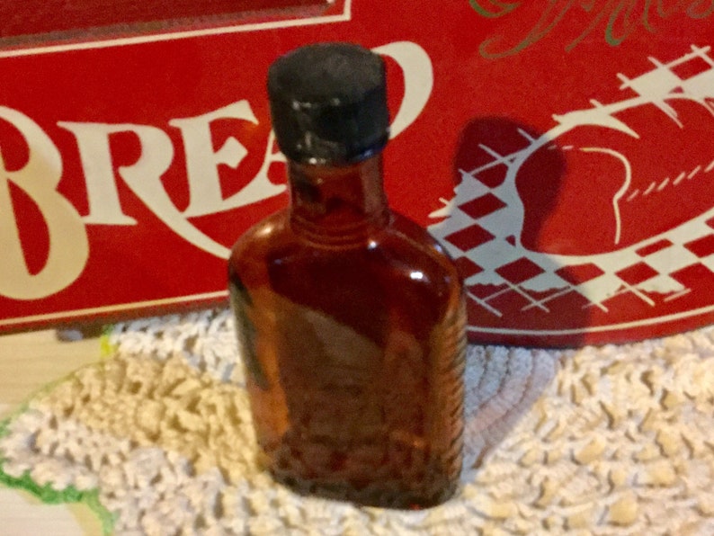 May include: A small, amber-colored glass bottle with a black cap. The bottle is sitting on a white crocheted doily.