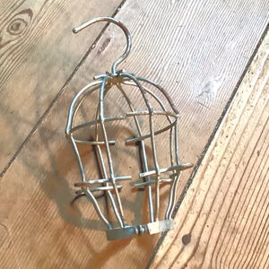 May include: A vintage, cage-style light fixture with a hook for hanging. The metal frame is a tarnished brass color, and the cage is designed to enclose a lightbulb. The fixture is resting on a wooden surface.