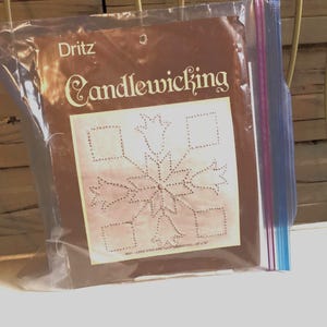 May include: A package of Dritz Candlewicking craft supplies. The brown package contains a cream-colored fabric with a snowflake design. The package is sealed in a clear plastic bag with a blue and purple zipper closure. The package is on a white surface.