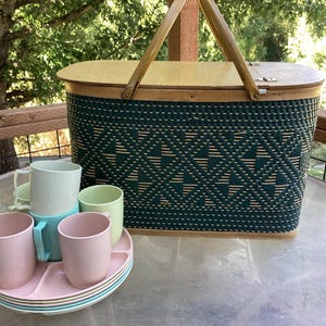 May include: A vintage picnic basket with a green woven design, wooden lid, and handle. Includes a set of pastel-colored cups and plates, perfect for outdoor dining. The basket measures approximately 18 inches long.
