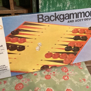 May include: A vintage Milton Bradley Backgammon game box with a yellow and black game board, red and black playing pieces, and two white dice. The box is blue and has the text "Backgammon and Acey-Deucy" printed on it.