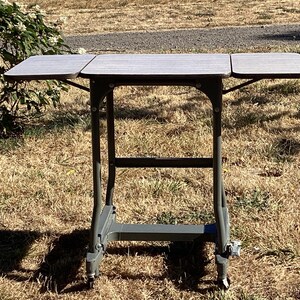 May include: A portable folding table with a gray metal frame and a rectangular top featuring two side leaves that extend outward. The table is supported by four legs with small wheels at the base, allowing for mobility and compact storage when folded.