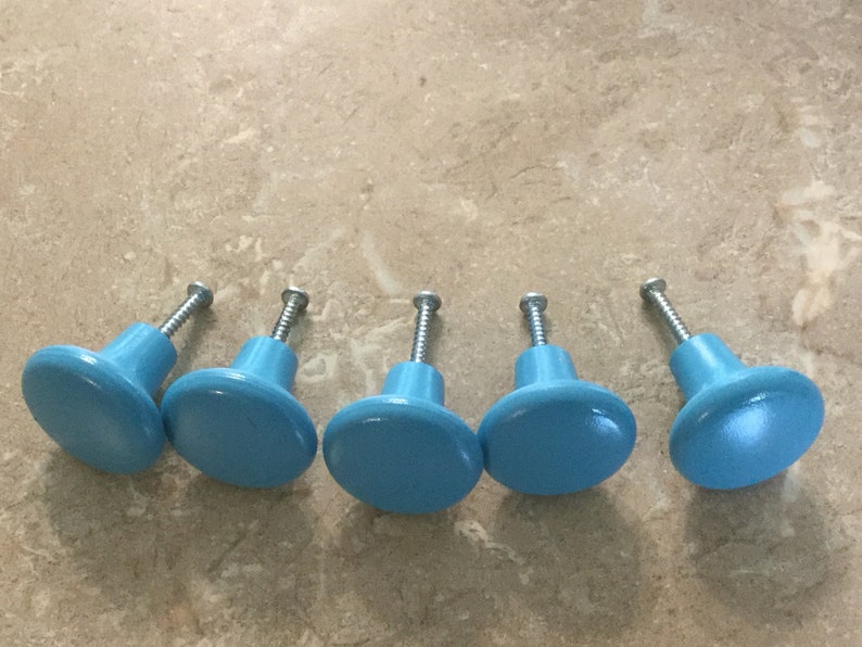 K&ouml;nnte beinhalten: F&uuml;nf hellblaue Schrankkn&ouml;pfe mit silbernen Schrauben. Die Kn&ouml;pfe sind rund und haben eine leicht erhabene Mitte.