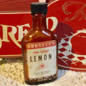 May include: A small amber glass bottle with a black cap. The bottle is labeled "Burnett's Pure Extract Lemon".