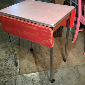 May include: A vintage red and pink drop-leaf table with a light pink tabletop and a red drop-leaf. The table has chrome legs and measures approximately 30 inches long when the leaves are down. The table is on wheels.