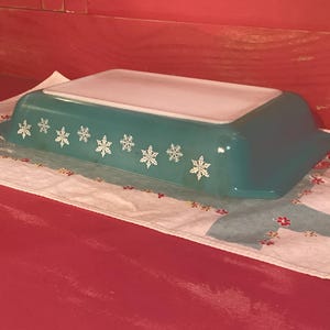 May include: A turquoise rectangular Pyrex baking dish with a white lid. The dish is decorated with white snowflake designs along the side. It rests on a floral patterned cloth.