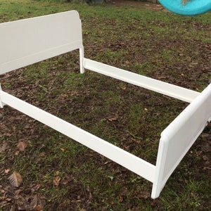 May include: White wooden bed frame with a headboard and footboard. The frame is on a grassy lawn with brown leaves.