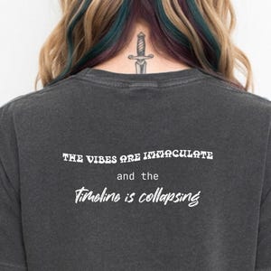 May include: Dark gray t-shirt with the text "THE VIBES ARE IMMACULATE and the Timeline is collapsing" in white. The shirt has short sleeves and a crew neck. The shirt is made of a soft, comfortable material.