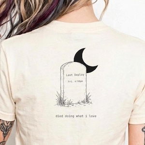 May include: Cream-colored t-shirt featuring a tombstone graphic with a crescent moon. The tombstone reads "Last Deploy" and "Fri. 4:58pm" with the phrase "died doing what I love" below. The shirt is a casual wear item.