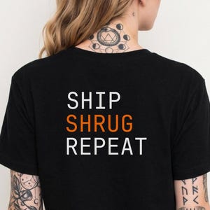 May include: Black t-shirt with the words "SHIP SHURG REPEAT" printed in white and orange. The shirt is a crew neck and has short sleeves. The text is centered on the back of the shirt.