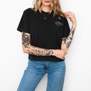 Puede incluir: Camiseta negra con un gr&aacute;fico blanco de una mano de esqueleto sosteniendo un cigarrillo, con el texto "-slow vibes" debajo. El modelo lleva vaqueros azules. El modelo tiene varios tatuajes en los brazos.