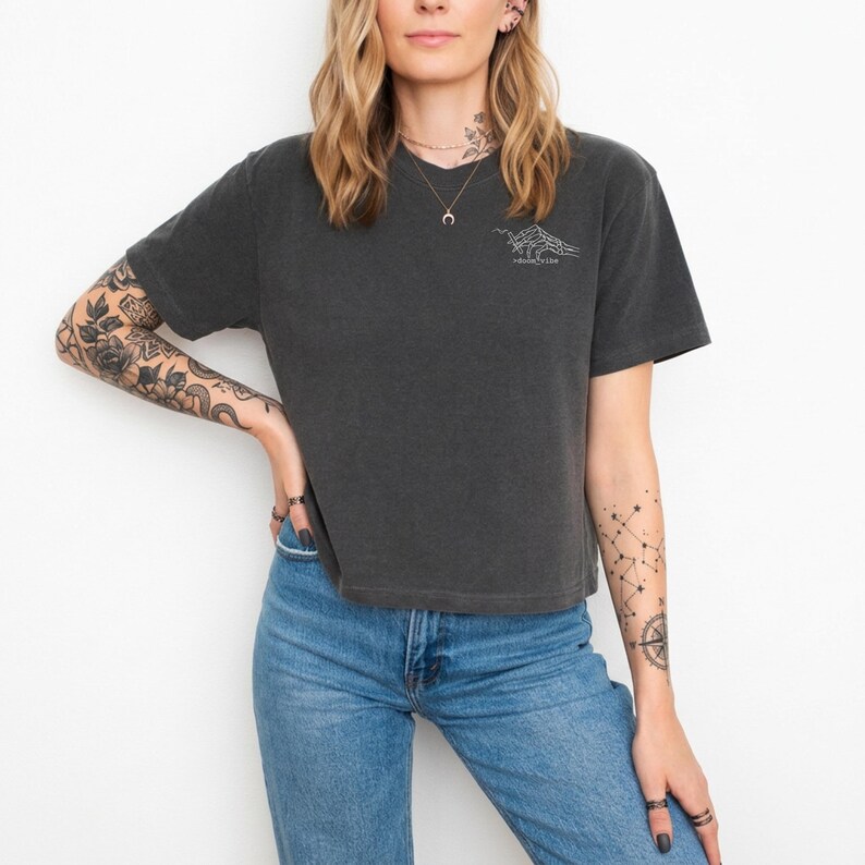 Puede incluir: Camiseta gris oscuro con cuello en V y mangas cortas. La camiseta presenta un peque&ntilde;o gr&aacute;fico blanco de una cordillera y el texto "wilderness love". La modelo lleva vaqueros azules.