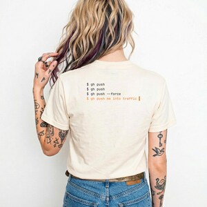 Puede incluir: Camiseta color crema con texto negro y naranja. El texto dice "$ gh push", "$ gh push --force" y "$ gh push me into traffic". La camiseta se combina con vaqueros azules.