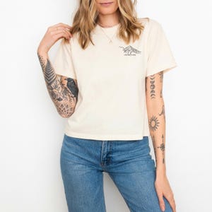 Puede incluir: Camiseta color crema con un gr&aacute;fico de monta&ntilde;a negro y el texto "desert vibe". La camiseta de manga corta se combina con unos vaqueros azules. La imagen muestra a una persona que lleva la camiseta.