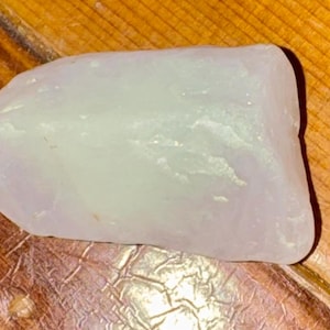 May include: A pale, translucent, irregularly shaped stone with a slightly rough texture. The stone is a soft, light lavender color and sits on a wooden surface with visible grain.