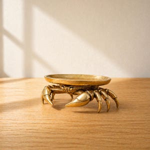 Puede incluir: Un cuenco para baratijas con forma de cangrejo de color dorado. El cangrejo tiene pinzas y patas detalladas, que sostienen un plato redondo y poco profundo. El plato es de metal y está sobre una superficie de madera.