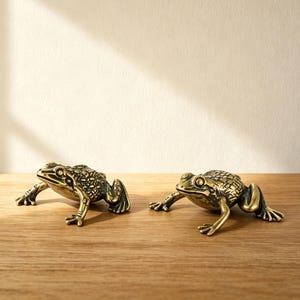May include: Two brass frog figurines with detailed textures are displayed on a wooden surface. The frogs are positioned side-by-side, facing forward, and are illuminated by natural light. The figurines have a vintage aesthetic.