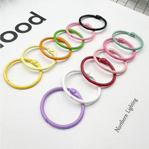 May include: A selection of brightly coloured metal binder rings in shades such as yellow, orange, green, pink, red, white, purple, and black. Each ring features a hinged opening. The rings are displayed on a white surface.