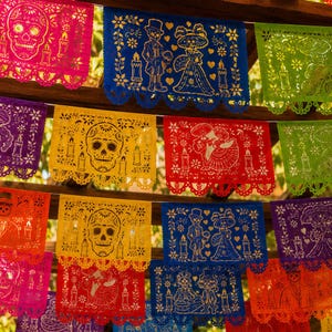 May include: A collection of colorful papel picado banners featuring intricate cut-out designs of skulls, skeletons in formal attire, and candles. The rectangular sheets are made of thin paper in shades of pink, blue, yellow, red, purple, and green.
