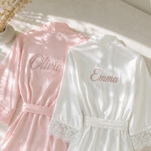 Puede incluir: Una bata de color rosa claro con el nombre Olivia bordado en cursiva y una bata blanca con el nombre Emma bordado en cursiva. Ambas batas cuentan con mangas largas con un borde de encaje ancho en los puños y un cinturón a juego.