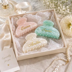 以下が含まれることがあります： パステルピンク、ミントグリーン、クリーム色のマーブル模様のヘアクリップ3点。それぞれにEmma、Sophie、Ameliaとゴールドの筆記体で名入れされています。クリップは白い薄紙が敷かれた正方形のギフトボックスに入っています。
