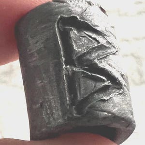 Könnte beinhalten: Ein dunkelgraues, zylindrisches Objekt mit geschnitztem Relief. Das Relief scheint eine stilisierte Figur oder ein Symbol darzustellen. Das Objekt wird zwischen zwei Fingern gehalten, was auf seine geringe Größe schließen lässt. Die Oberfläche hat ein strukturiertes, verwittertes Aussehen.