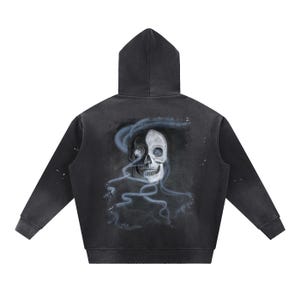 Puede incluir: Sudadera con capucha negra con acabado desgastado. La parte trasera presenta una ilustración detallada de una calavera con un diseño de media cara, acentuada con elementos azules en forma de humo. La sudadera tiene capucha y mangas largas.