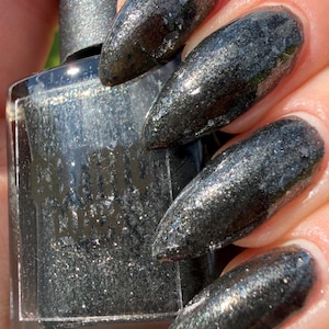May include: A rectangular glass bottle of nail polish with a black cap, featuring a dark charcoal gray liquid with fine silver glitter and metallic flakes. The bottle has the text "CLIONA LUNA" printed in black on the front surface.