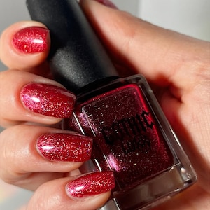 May include: A bottle of red nail polish with a black cap is held in a hand. The nails are painted with the same sparkling red polish. The bottle is square with a silver base.