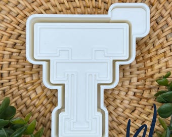 May include: A white plastic cookie cutter shaped like the letter T with a blocky, collegiate-style font design. The cutter features a raised inner edge for detailed impressions and a wider outer rim for cutting through dough cleanly and efficiently.