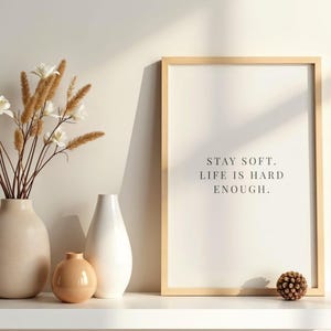 Puede incluir: Una impresión enmarcada con el texto "STAY SOFT. LIFE IS HARD ENOUGH." se encuentra en una estantería blanca, junto a una colección de jarrones y flores secas. El marco es de madera clara y los jarrones son de tonos neutros.