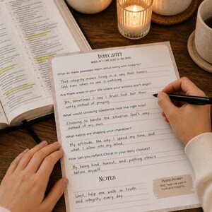 May include: A person writing on a lined journal page with the word "INTEGRITY" at the top. The page is part of a prayer prompt journal with questions about integrity. A Bible, candles, and a cup of tea are in the background.