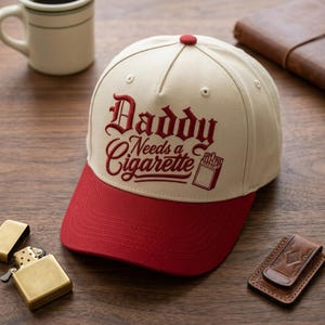 May include: A cream-colored baseball cap with a red brim and a red button on top. The front features embroidered dark red text that reads Daddy Needs a Cigarette, accompanied by a small graphic of a cigarette pack.