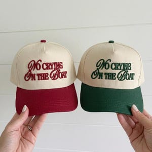 May include: Two baseball caps with cream-colored crowns and solid-colored brims. One cap features a deep red brim and matching red embroidered text that reads No Crying On The Boat. The other cap has a dark green brim and matching green text.