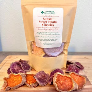 May include: A brown paper bag of Sunset Sweet Potato Chewies dog treats. The bag has a white label with text and a green clover logo. The treats are purple and orange sweet potato slices, some are outside the bag on a wooden board.