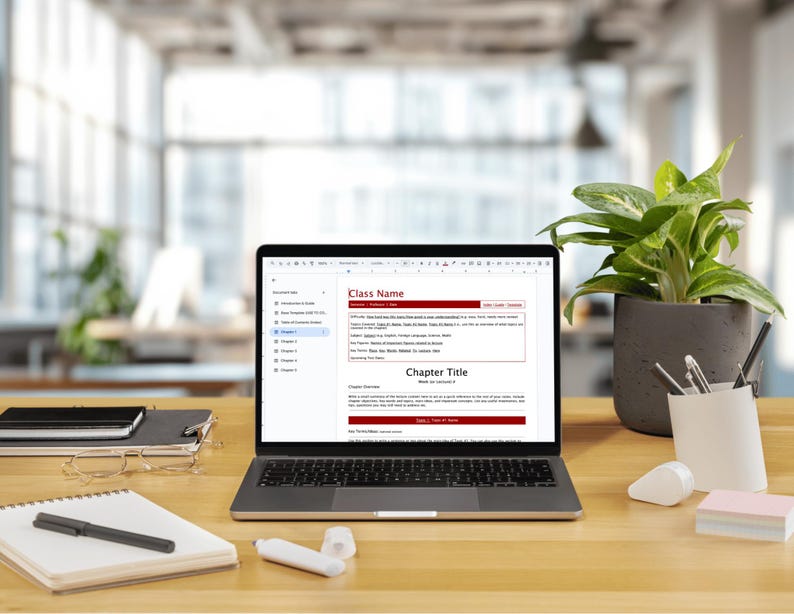 May include: A silver laptop displaying a document with the text Class Name and Chapter Title on its screen. The laptop sits on a wooden desk alongside a spiral notebook, a black pen, a white correction fluid, a potted green plant, and a pen holder.