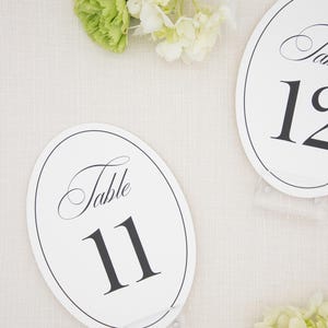 May include: Two oval white table number cards featuring black text that reads Table 11 and Table 12. Each card has a thin black border and is displayed on a clear acrylic stand, accented by clusters of light green and white hydrangea flowers.