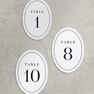 May include: Three oval-shaped white table number cards featuring a thin black double-line border. Each card displays the word TABLE in black capital letters above the large black numerals 1, 8, and 10, printed in a classic serif font style.