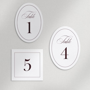 May include: White table number cards with burgundy text. Two oval cards with the word "Table" and numbers 1 and 4. A square card with the number 5. Elegant design for weddings or events.