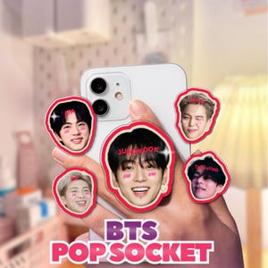 Puede incluir: Un teléfono inteligente blanco sostenido en una mano, con cinco pop sockets con bordes rojos que presentan imágenes de miembros de BTS. Los pop sockets tienen varias formas y tamaños. El texto "BTS POPSOCKET" se muestra en rosa y morado.