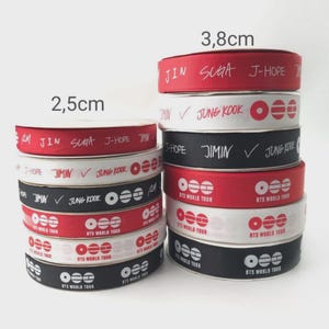 Könnte beinhalten: Zwei Stapel Ripsband in Rot, Weiß und Schwarz. Der linke Stapel ist 2,5 cm breit, der rechte 3,8 cm. Die Bänder sind mit Texten bedruckt, darunter Namen wie Jin, Suga, J-Hope, Jimin und Jung Kook sowie das Logo der BTS World Tour.