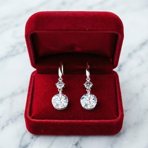 May include: Silver dangle earrings with clear, round gemstones, presented in a red velvet box. The earrings feature a small gemstone above a larger, round, faceted stone. The background is a marble surface. The earrings are a classic jewellery item.