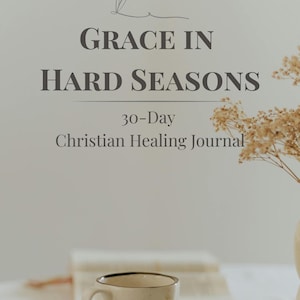 May include: A beige ceramic mug with a dark brown rim and handle sits on a saucer. The image features a journal with the text "GRACE IN HARD SEASONS" and "30-Day Christian Healing Journal". Dried flowers and a pen are also visible.
